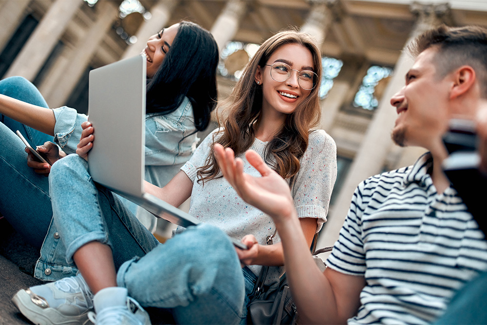 Three college students sitting cross-legged, smiling and laughing. Female in the center is holding a laptop computer